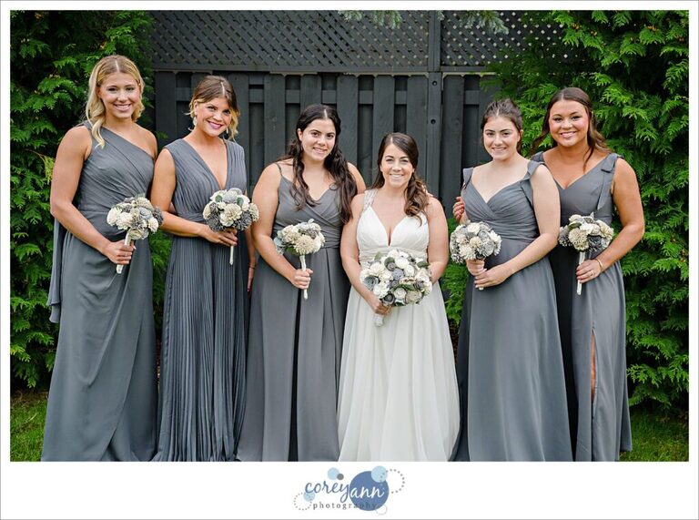 Group of women standing together center wearing a white bridal gown and the others in grey toned bridesmaid dresses