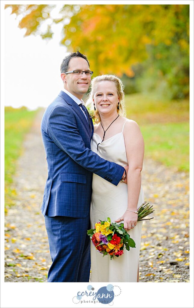 A bride in a slip dress poses with her groom in a blue suit with a bright floral bouquet.