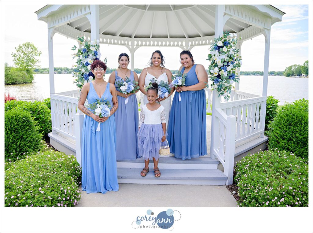 Bride in a long white dress with spaghetti straps and bridesmaids wearing blue dresses in a white gazebo on a lake in june in Akron Ohio