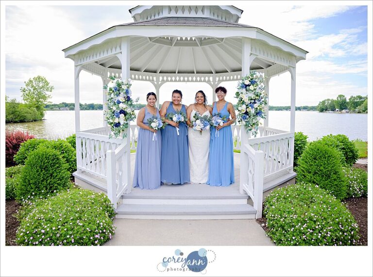 Bride in a long white dress with spaghetti straps and bridesmaids wearing blue dresses in a white gazebo on a lake in june in Akron Ohio