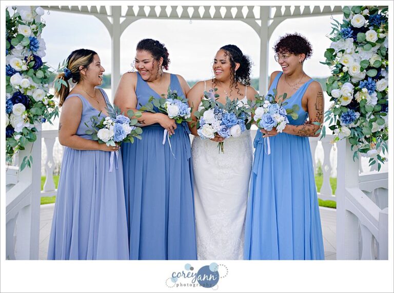 Bride in a long white dress with spaghetti straps and bridesmaids wearing blue dresses in a white gazebo on a lake in june in Akron Ohio