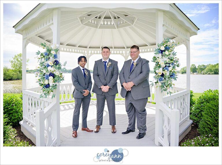 A man who is the groom standing between his two groomsman all wearing light grey suits inside a white gazebo on a sunny June day that is decorated for a wedding in Akron Ohio