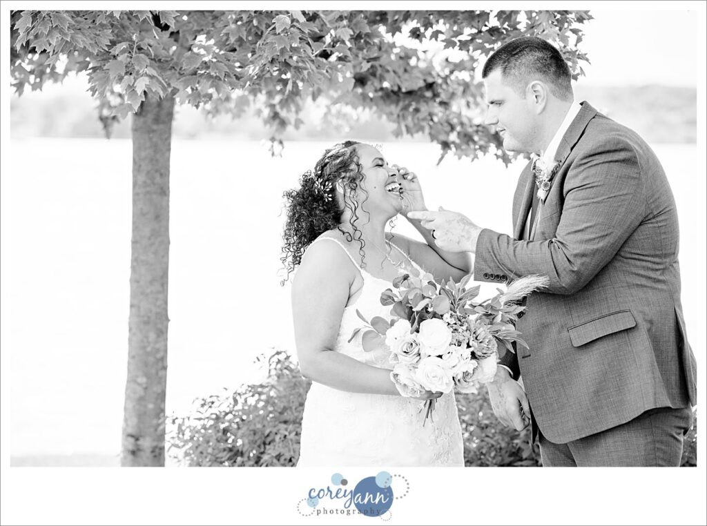 Black and white portrait of a bride in a white dress holding a floral bouquet looking at her groom in a suit in front of a tree and a lake in Akron Ohio