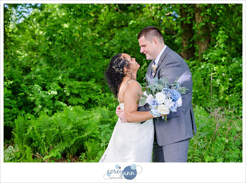 Bride and groom looking at each other and smiling in front of green trees on a sunny Ohio June day 