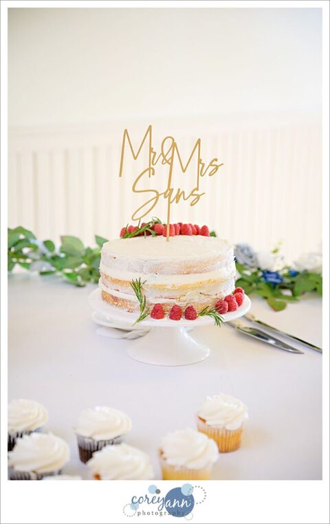 A simple three layered wedding cake that is in the nearly naked style with barely any frosting on a white pedestal with a few berries placed around it. On top of the cake is a Mr and Mrs cake topper in gold with hand written font. The cake is on a table in front of a white background. 