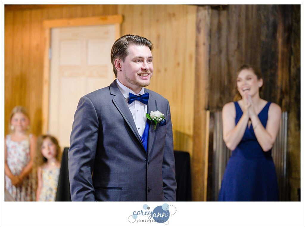 Groom wearing a charcoal tux with blue bow tie and vest smiling when seeing his bride for the first time at Amy's Rustic Event Center in Valley City, Ohio
