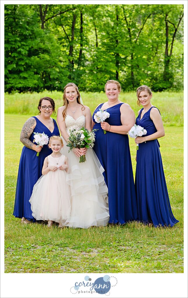 A bride and her 3 bridesmaids and flower girl posing before the wedding ceremony at Amy's Rustic Event Center in Valley City, Ohio in June. The bridesmaids are wearing long blue gowns with thicker stank straps and the flower girl is wearing a light pink dress that echos the style of the bridal gown. 