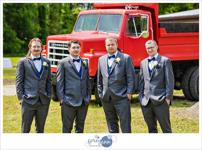 A groom and three groomsman posing before the wedding ceremony at Amy's Rustic Event Center in Valley City, Ohio in June. They are standing in front of a red dump truck all wearing charcoal suits with navy vests and bow ties. 