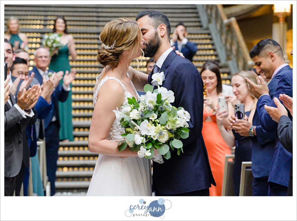 Bride carrying a bouquet of white and green kissing a groom in a tailored suit after their ceremony at the Hyatt Regency Cleveland at The Arcade in Ohio