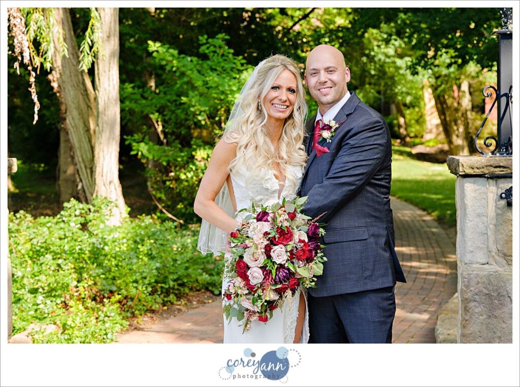 A bride wearing a white dress with a sweetheart neckline standing next to a groom in a dark gray suit, she is holding a large bouquet of flowers in light pink and red and they are standing in front of a path through Gervasi Vineyard on a sunny day in Canton Ohio for their July wedding