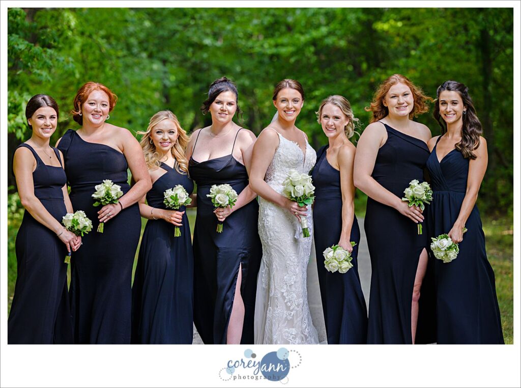 Bridesmaids wearing various black gowns pose alongside the bride while holding white floral bouquets at Huntington Reservation after wedding in Ohio