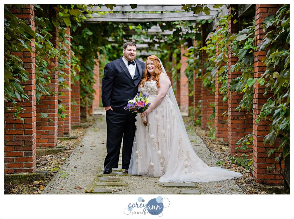 Bride wearing a floral WToo dress with a groom wearing a traditional black tux posing in the arbor at Stan Hywet in Akron Ohio after their wedding. 