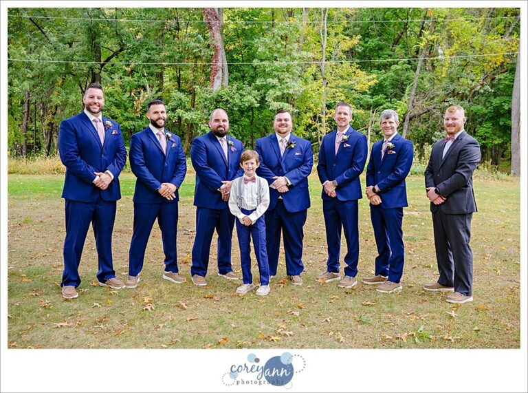Groom and groomsman wearing blue suits with pink ties posing near trees at Tudor House on Portage Lakes