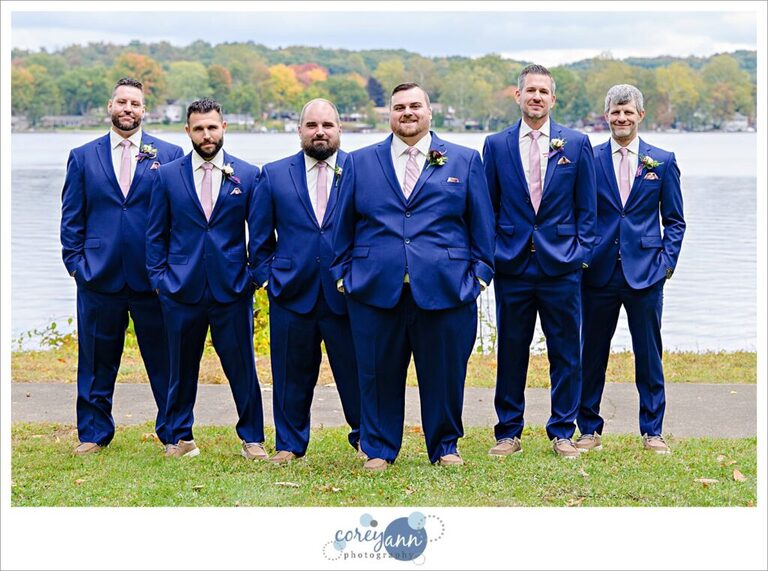 Groom and groomsman outside wearing blue suits with pink ties and posing in a v formation near a lake in Akron Ohio