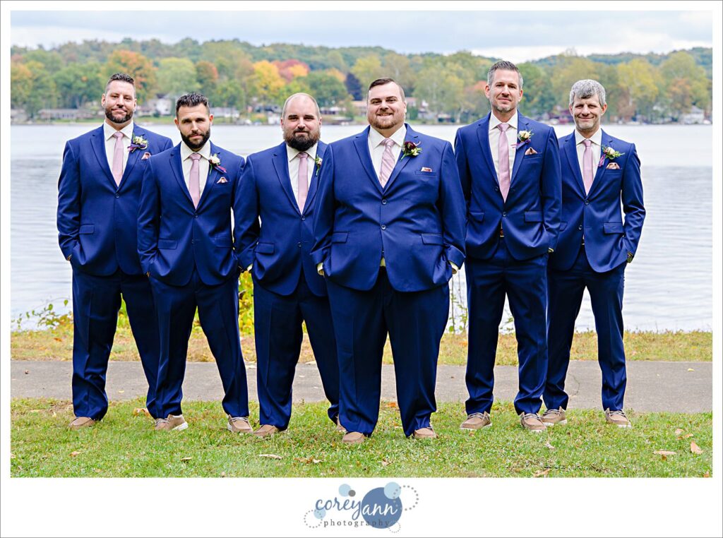 Groom and groomsman outside wearing blue suits with pink ties and posing in a v formation near a lake in Akron Ohio