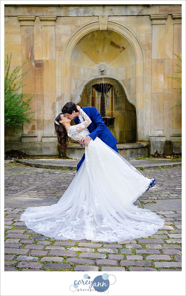 Groom dipping and kissing his bride in front of the fountain with her train spread out at the Italian Cultural Gardens in Cleveland before their wedding in Ohio